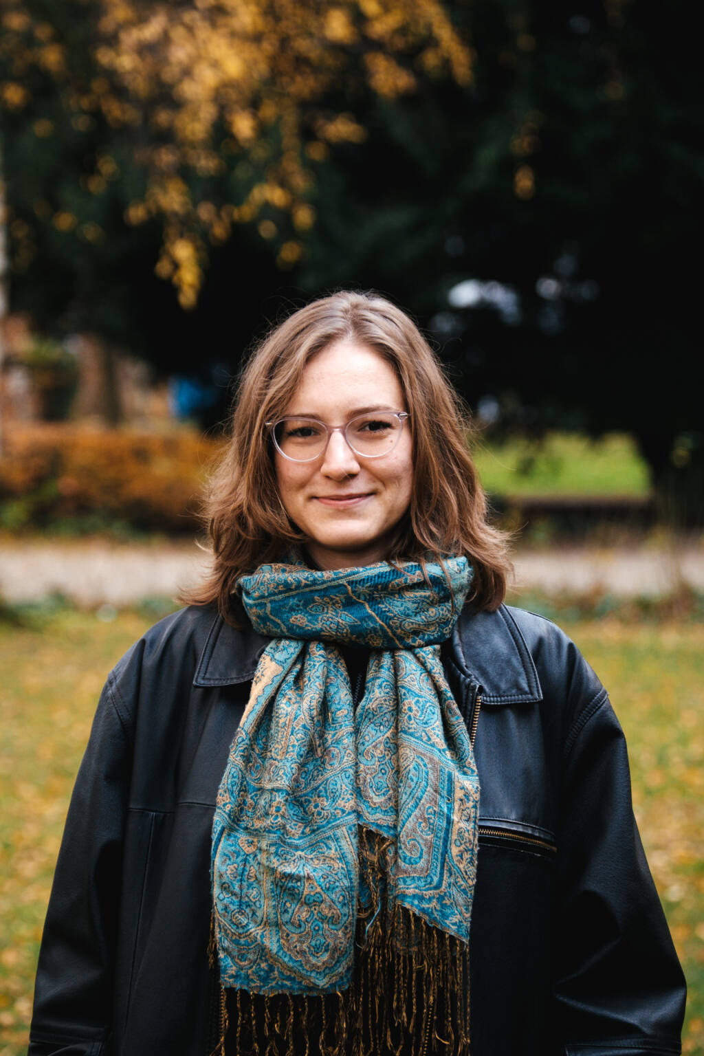 Porträt von Lena Hofmann im Freien vor einem herbstlichen Hintergrund. Sie blickt lächelnd in die Kamera, trägt eine Brille, eine schwarze Lederjacke und einen gemusterten, blau-goldenen Schal.